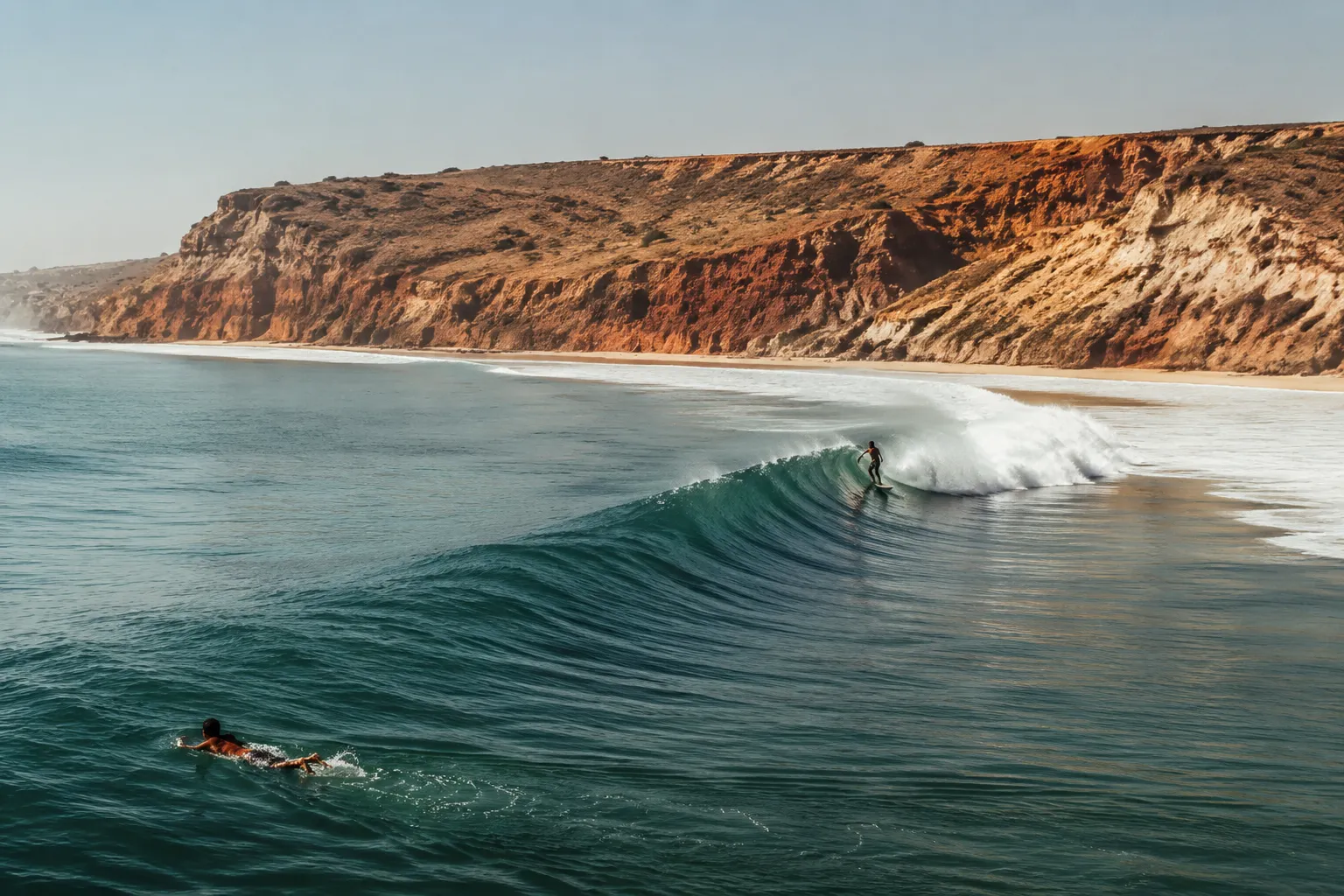 A surfer riding the long left-hand point break at Cabo Ledo, along a golden bay backed by red-earth cliffs south of Luanda