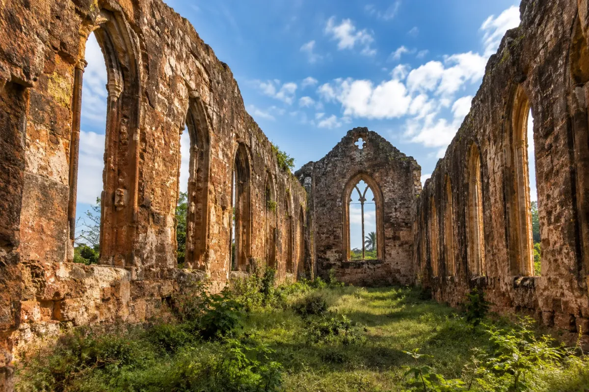 Roofless nave of a ruined stone church in tropical Africa, empty arched windows and grass growing across the floor